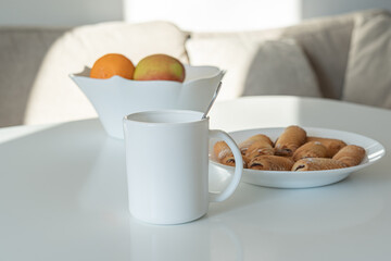 A white cup stands on a bright table. In the background are fruits and cookies.