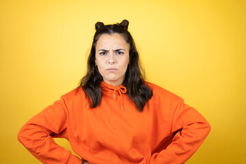 Young beautiful woman wearing sweatshirt over isolated yellow background skeptic and nervous, disapproving expression on face with arms in waist