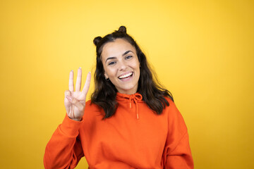 Young beautiful woman wearing sweatshirt over isolated yellow background showing and pointing up with fingers number three while smiling confident and happy