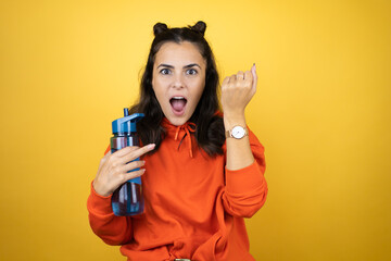 Young beautiful woman wearing sweatshirt holding a water bottle over isolated yellow background surprised and showing her watch because it's late