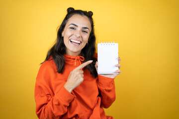 Naklejka premium Young beautiful woman wearing sweatshirt over isolated yellow background smiling, surprised and pointing blank notebook in her hand