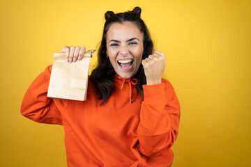 Young beautiful woman wearing sweatshirt over isolated yellow background holding a paper bag very happy and excited making winner gesture with raised arms, smiling and screaming for success