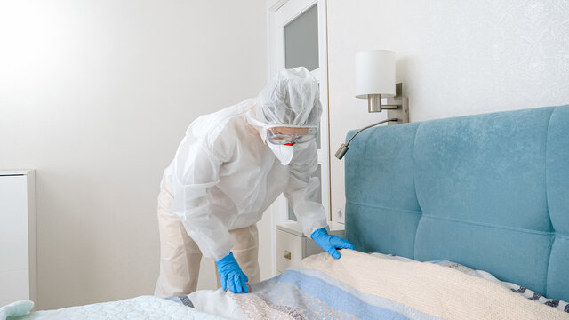 Young housewife in protective biohazard suit making up bed during covid-19 pandemic and lockdown - Powered by Adobe