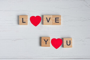 Top view of the word love you laid out from square wooden tiles and smal red hearts on old white wooden background. Concept of valentine day.