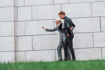 Walking on grass near wall. Young guy with senior man in elegant clothes is outdoors together. Conception of business