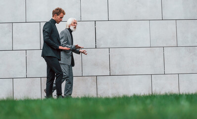 Walking on grass near wall. Young guy with senior man in elegant clothes is outdoors together. Conception of business