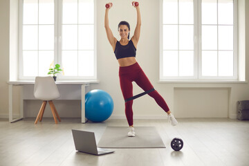 Active healthy lifestyle, training at home concept. Smiling young woman athlete in sportswear doing workout with fitness gum and dumbbels on fitness mat at home online during quarantine