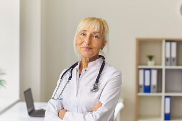 Smiling elderly woman doctor thetapist in white medical uniform standing and looking at camera in clinic office over desk with laptop at background. Medicare, healthcare, therapist concept