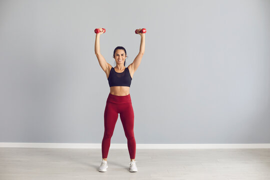 Lifestyle. Portrait of a beautiful athletic woman standing with raised dumbbells in her hands and showing off her toned athletic body. Woman in sports uniform posing on a background of gray wall.
