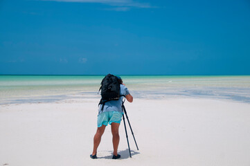 A Photographer with his camera takes landscape photographs at low tide on the island of Holbox, Mexico. In the background the blue sky and a heron on the sea