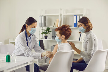 Vaccination and immunization for children concept. Child getting injection at the hospital and mom supporting him. Nurse or doctor in medical face mask giving flu or Covid-19 vaccine to little boy
