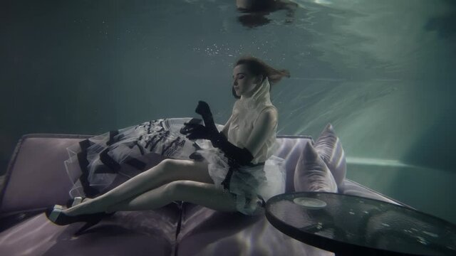 funny and eccentric woman is sitting on couch underwater and playing with floating cup