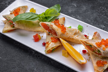 Banquet serving of pancakes with red caviar and salmon, on a white rectangular plate, with basil leaves and an orange slice