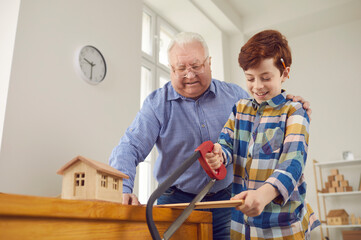 Little kid and his grandfather building toy houses and having fun in workshop. Smiling boy sawing...