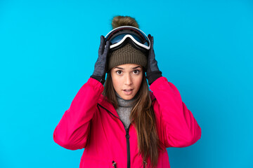 Teenager skier girl with snowboarding glasses over isolated blue background with surprise facial expression