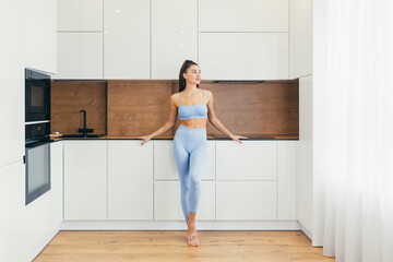 Fototapeta na wymiar Strong and Beautiful Athletic Fitness Girl in Sportswear is Doing Exercises in Her Bright and Spacious Kithen with Minimalistic Interior