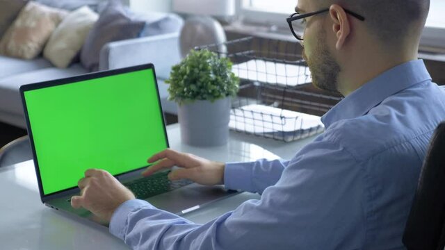Rear View Of A Green Monitor Screen and A Young Man Working On An Online Learning Project On A Computer Using A Laptop In A Bright Cozy Room At Home