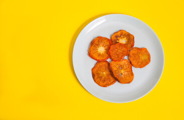 Persimmon chips lie on a porcelain plate on a yellow background. Copy space.