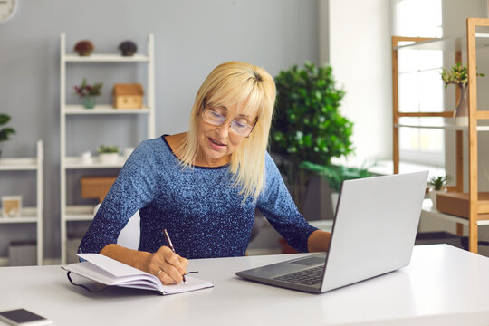 Senior Woman Making Notes In Notebook While Looking At Business Training Or Online Webinar On Laptop. Concept Of Remote Work Or Social Distance Learning At Home.