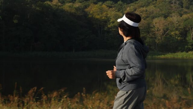 Side view, medium shot of senior Asian woman wearing gray sweater and headband, running at the road of the reservoir to workout in the morning, Active lifestyle and vitality concept.