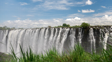 Die Viktoriafälle zwischen Simbabwe und Sambia mit Gras im Vordergrund und blauen Himmel im Hintergrund