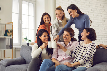 Obraz premium Group of happy young women having fun and enjoying time together. Smiling friends or colleagues sitting on sofa at home or in office, laughing at funny joke, clinking cups and drinking coffee or tea