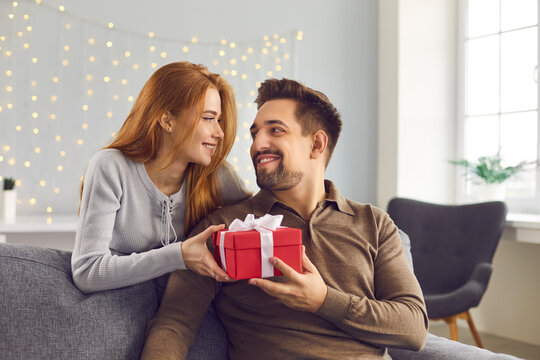 Beautiful young couple at home. Romantic woman stands behind a man sitting on a sofa and hands him a holiday present. Concept of gifts for birthdays, Christmas, Valentine's Day and Men's Day.