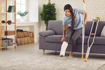 Young woman with broken leg in plaster cast trying to stand up from sofa and walk with crutches in...