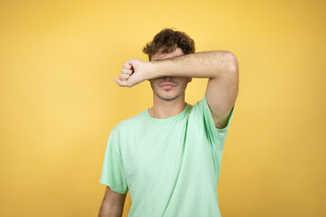 Handsome man wearing a green casual t-shirt over yellow background covering eyes with arm smiling cheerful and funny. Blind concept.