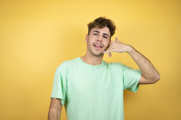 Handsome man wearing a green casual t-shirt over yellow background smiling doing phone gesture with hand and fingers like talking on the telephone