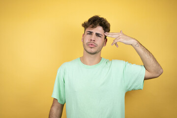 Handsome man wearing a green casual t-shirt over yellow background Shooting and killing oneself pointing hand and fingers to head like gun, suicide gesture.