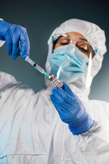 Close-up of female Scientist researcher face, drawing the liquid from Covid-19 vaccine bottle with a syringe in a research medical lab. Surgical mask and gloves, and PPE suit. Coronavirus Pandemic. 