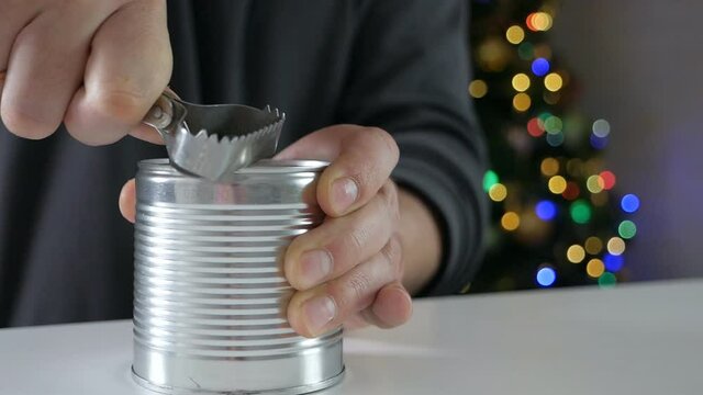 Close-up shot of strong male hands with opener making an incision on the lid of a metal can in front of the christmas tree
