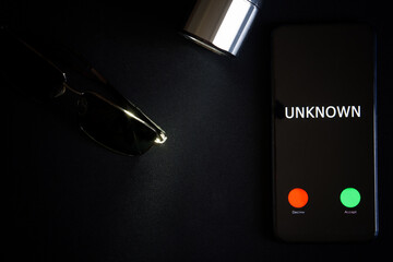 A smartphone with an unknown call lies on a black surface next to sunglasses and a flashlight. The problem of anonymity and espionage with modern gadgets