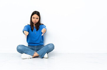 Young mixed race woman sitting on the floor isolated on white background showing thumb down with two hands