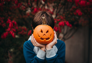 Young boy holding a mini jack-o-lantern over his face outdoors.