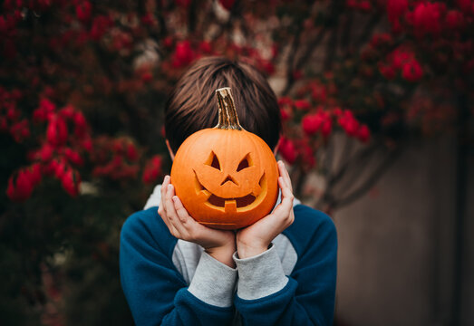 Young boy holding a mini jack-o-lantern over his face outdoors.