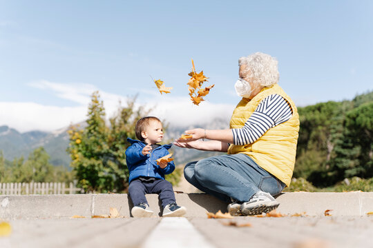 Grandmother wearing medical mask playing with leaves with her little grandson in a park