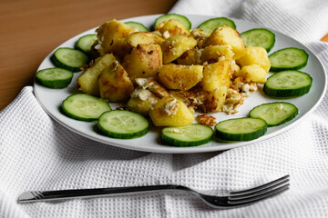 Fried potato slices in a beaten egg with protein, around a circle of green juicy fresh cucumber. Dish on a white plate on a wooden table