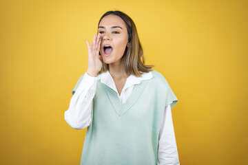Pretty young woman standing over yellow background shouting and screaming loud to side with hands on mouth