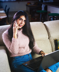 Beautiful young girl on beije sofa interacting with laptop and cellphone using social media while studying and working from home thanks to internet.