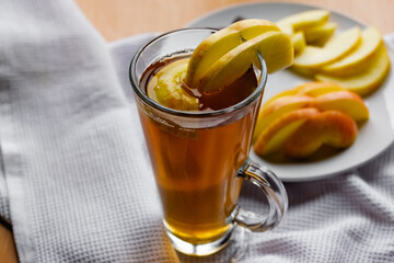 Apple tea in a transparent glass with a handle, the drink is decorated with slices of fresh apple with a yellow peel, next to the plate of sliced fruit and chocolate, everything is on the table on a w