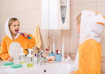 Five year old girl wearing orange pyjamas brushes her teeth with electric toothbrush in a bathroom