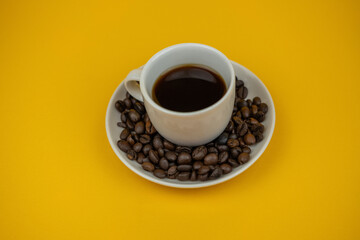 A nice photo of a little cup of coffee surrounded by some coffee beans in a plate on a yellow background
