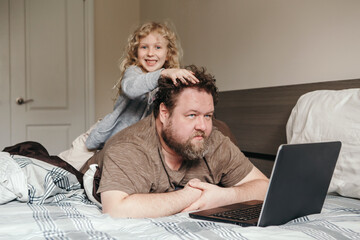 Work from home with kids children. Father working on laptop in bedroom with child daughter on his back. Funny candid family moments. New normal during coronavirus quarantine lockdown. - 403865733