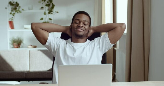Portrait of smiling contented African American black man in white T-shirt sits at table with his hands folded behind his head resting enjoying youthful life and good news rejoices dreaming takes break