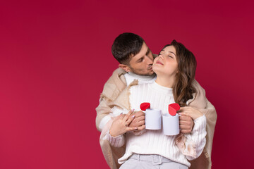 A loving couple wrapped in a warm blanket holds hot tea cups have a lovely time. Red background. St. Valentine's Day.