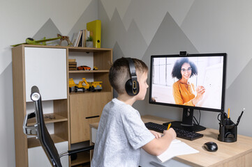 Above shoulder shot of a cute schoolkid watching online video classes sitting on self isolation at home, a smiling female teacher on the screen. E-learning, distance education