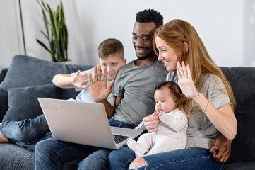 Multiracial family sitting on the comfortable sofa and using laptop for video call, spouses and their kids waving in webcam and smile. Virtual meeting, online talking