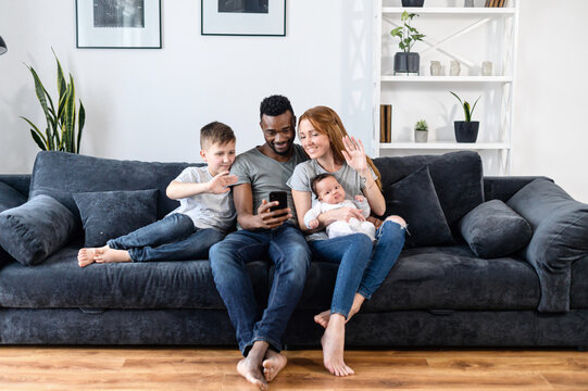 A diverse loving family of four using smartphone for video connection with family, friends, grandparents. A multiracial parents and their two childs on the sofa waving hello into webcam